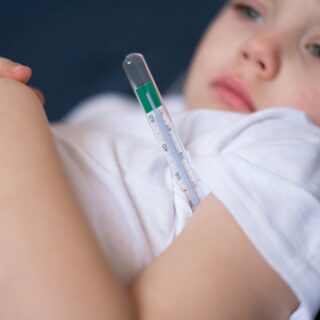 صبي صغير يرتدي كنزة بيضاء ويتمدد في السرير ويضع تحت ابطه ميزان لقياس درجة حرارة الجسم