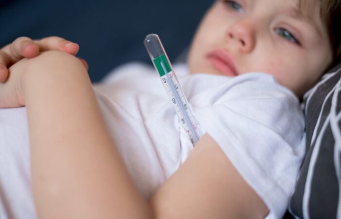 صبي صغير يرتدي كنزة بيضاء ويتمدد في السرير ويضع تحت ابطه ميزان لقياس درجة حرارة الجسم