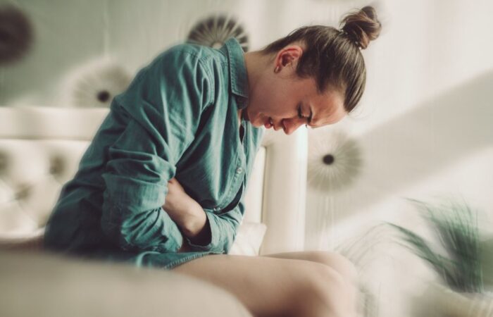 امرأة ترتدي كنزة خضراء تجلس على السرير وتضع يديها على بطنها وتخفض رأسها للأسفل بسبب شعورها بالألم