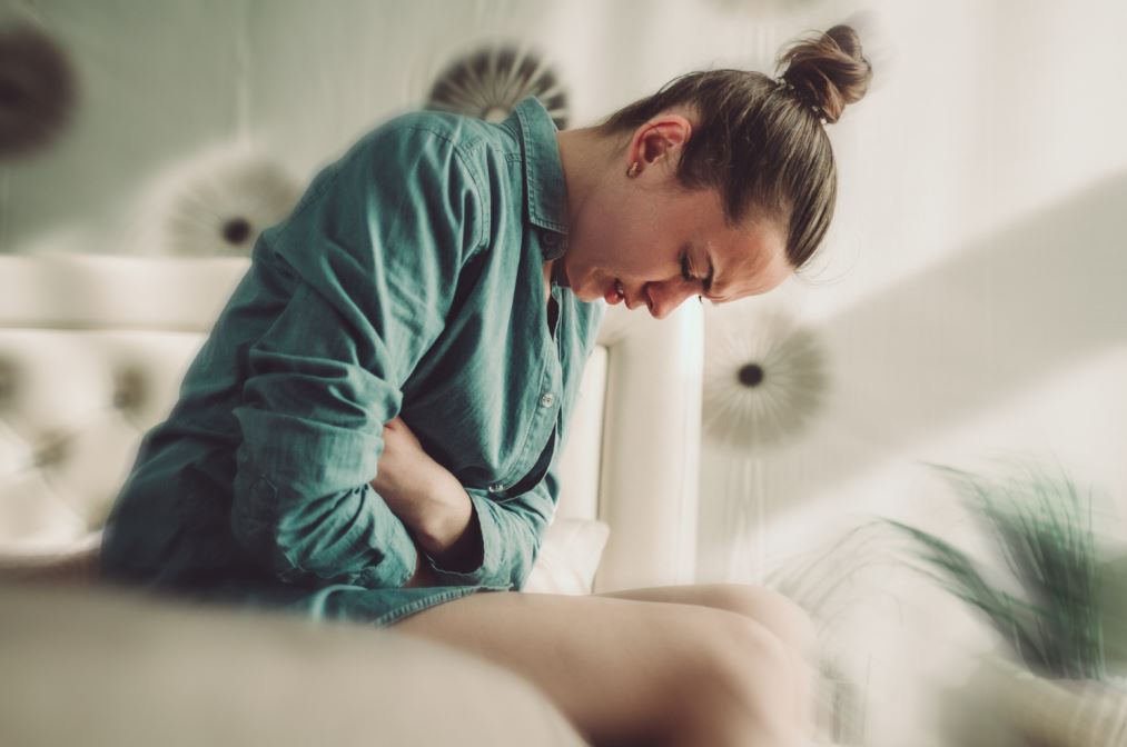 امرأة تجلس على السرير وتضع يديها على بطنها وتخفض رأسها للأسفل بسبب شعورها بالألم