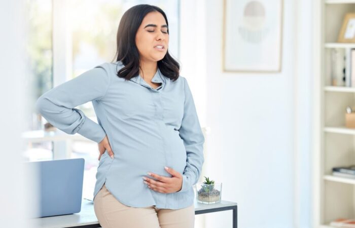 امرأة حامل مغمضة العينين تلبس قميص أزرق وتضع يد خلف ظهرها واليد الآخرى أسفل بطنها