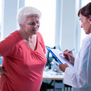 طبيبة تلبس قميص أبيض تقوم باستجواب سيدة مُسنة تلبس كنزة حمراء تقف أمامها وتقوم هذه السيدة بوضع يديها خلف ظهرها