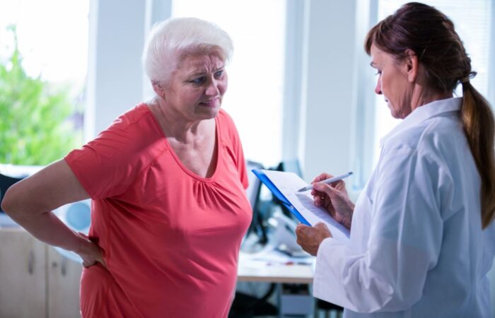 طبيبة تلبس قميص أبيض تقوم باستجواب سيدة مُسنة تلبس كنزة حمراء تقف أمامها وتقوم هذه السيدة بوضع يديها خلف ظهرها