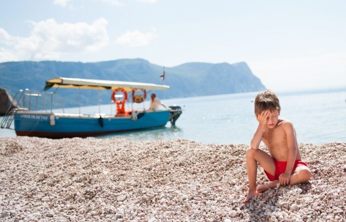 صبي صغير يجلس على الشاطئ أمام قارب صيد في وقت الظهر ويضع يده على جبينه