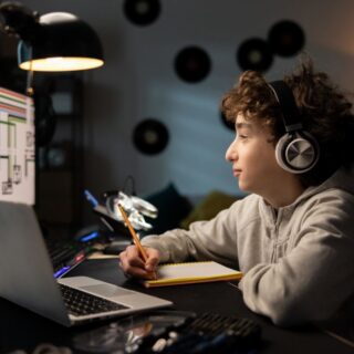 صبي يجلس أمام شاشة الكمبيوتر ويضع سماعات على رأسه ويمسك بيده قلم للكتابة على دفتر