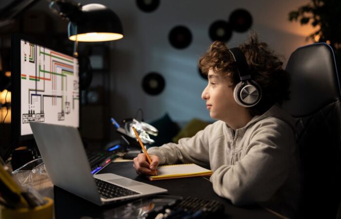 صبي يجلس أمام شاشة الكمبيوتر ويضع سماعات على رأسه ويمسك بيده قلم للكتابة على دفتر