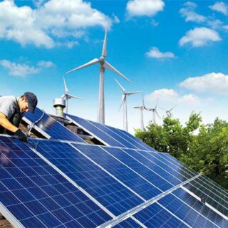 عامل بناء يقوم بتركيب الألواح الشمسية سوداء اللون على سطح منزل وخلف المنزل تظهر مزرعة لتوربينات الرياح