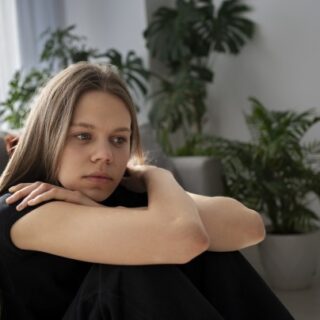 فتاة ترتدي كنزة سوداء تجلس على الأرض متشابكة الأيدي وتظهر علامات الحزن على وجهها