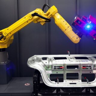 ذراع روبوت أصفر تحمل ماسحاً ضوئيا وتقوم بتصوير قطعة من هيكل سيارة داخل غرفة مظلمة