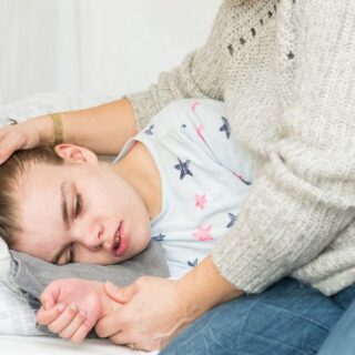 فتاة ممددة في السرير تظهر ملامح الخوف على وجهها ويقوم شخص يجلس بجانبها بوضع يده على رأسها ويمسك بيده الأخرى يدّ الفتاة