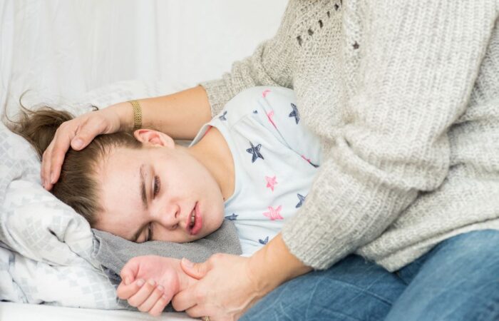 فتاة ممددة في السرير تظهر ملامح الخوف على وجهها ويقوم شخص يجلس بجانبها بوضع يده على رأسها ويمسك بيده الأخرى يدّ الفتاة