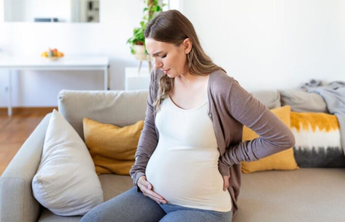 امرأة حامل تجلس على الأريكة وتضع يد على خصرها واليد الأخرى تضعها أسفل بطنها