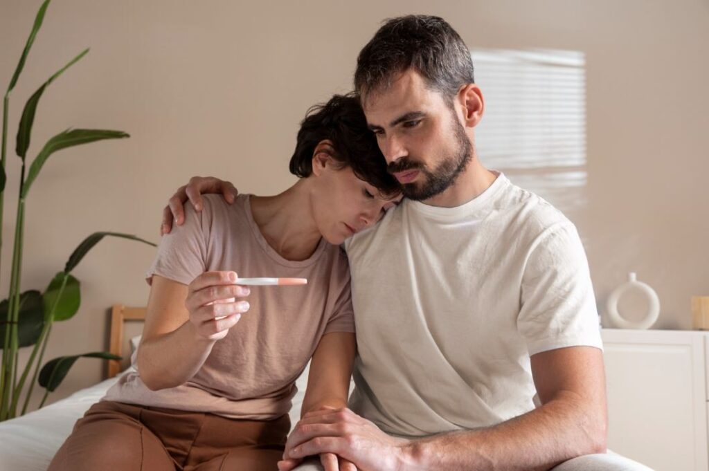 رجل بالغ يقوم بحضن امرأة تجلس بجانبه وهي تحمل بيدها جهاز لتحليل الحمل