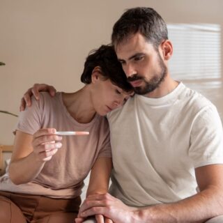 رجل بالغ يقوم بحضن امرأة تجلس بجانبه وهي تحمل بيدها جهاز لتحليل الحمل