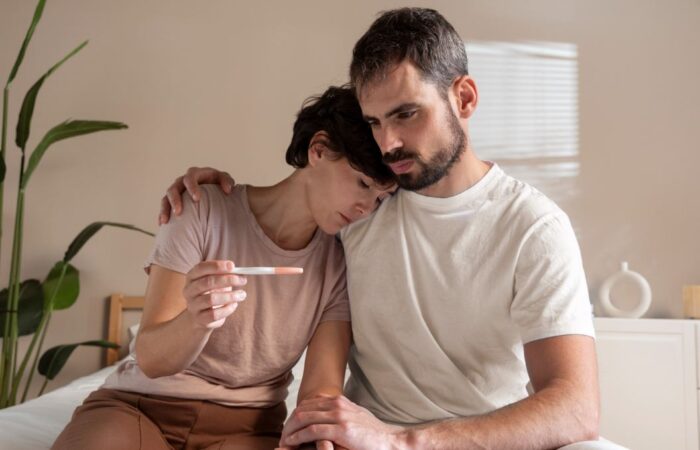 رجل بالغ يقوم بحضن امرأة تجلس بجانبه وهي تحمل بيدها جهاز لتحليل الحمل