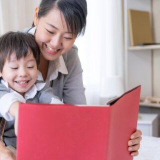 صبي صغير يقرأ كتاب لون غلافه أحمر وتقوم امرأة بحضنه من الخلف