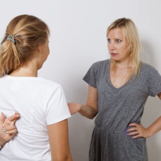 فتاة تقف أمام امرأة بالغة وتقوم هذه الفتاة بصنع إشارة خاصة بأصابع يديها التي تخفيها خلف ظهرها