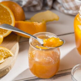 علبة زجاجية شفافة بداخلها مادة صفراء وعلى أحد العلب توجد ملعقة بها مادة صفراء وحول العلبة توجد العديد من قطع البرتقال