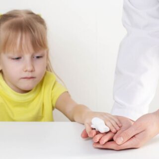 فتاة صغيرة ترتدي كنزة صفراء وفي إصبعها جهاز قياس و تقوم بوضع يدها على يد شخص يرتدي كنزة بيضاء جانبها