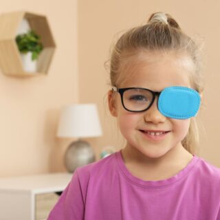 فتاة صغيرة ترتدي كنزة زهرية وتضع قطعة من القماش الأزرق على العدسة اليسارية لنظارتها الطبية