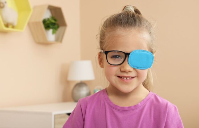 فتاة صغيرة ترتدي كنزة زهرية وتضع قطعة من القماش الأزرق على العدسة اليسارية لنظارتها الطبية