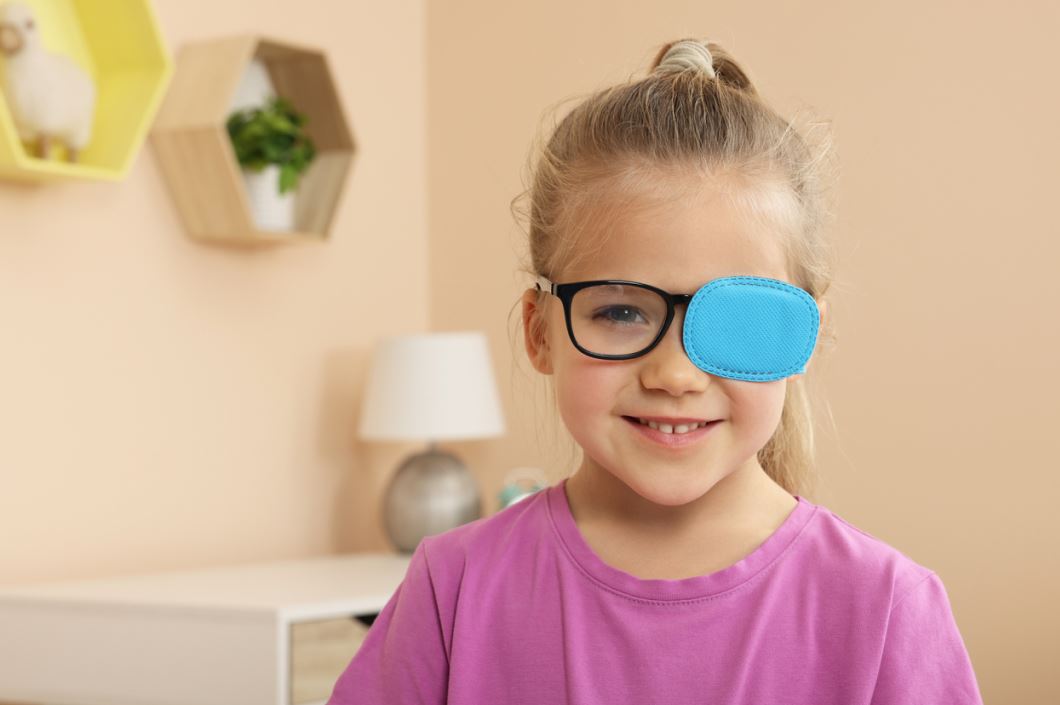 فتاة صغيرة ترتدي كنزة زهرية وتضع قطعة من القماش الأزرق على العدسة اليسارية لنظارتها الطبية
