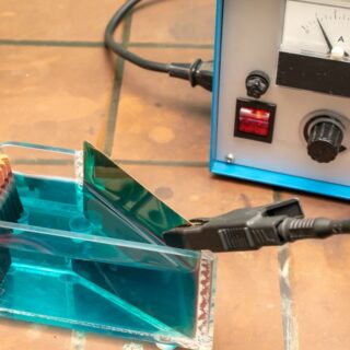 علبة شفافة تحتوي على سائل أزرق يوجد به قطعتين معدنيتين موصولتين بكبلان أسود وأحمر يخرجان من جهاز الكتروني عليه مفتاح دائري أسود كبير