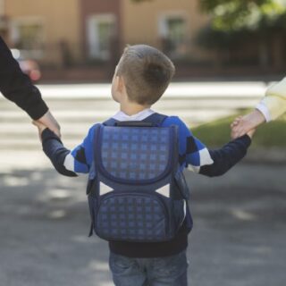 طفل يرتدي حقيبة مدرسية ويمسك يديّ رجل وامرأة على جانبيه
