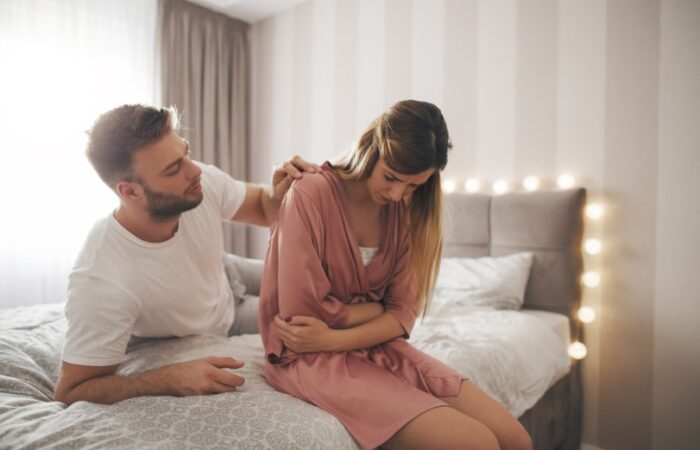 رجل ممد على السرير يقوم بوضع يده على كتف امرأة تجلس بجانبه تنحني للأمام وتضع يديها على بطنها