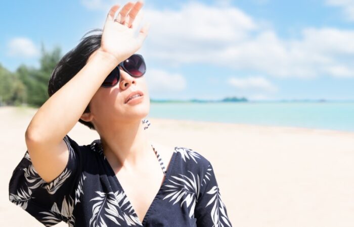 فتاة ترتدي قميص أسود ونظارات شمسية تسير على شاطئ البحر في يوم مشمس وتضع يدها على جبينها