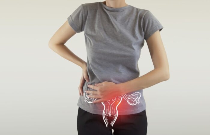 امرأة ترتدي كنزة رمادية تضع يد على خصرها واليد الأخرى أسفل بطنها حيث توجد رسمة لرحم الأنثى