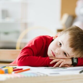 طفل يرتدي كنزة حمراء ويضع رأسه على الطاولة وأمامه أقلام ملونة