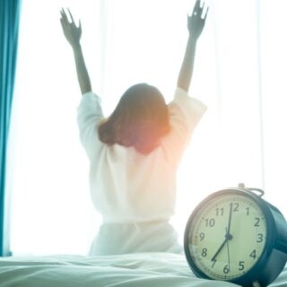 فتاة تجلس على السرير رافعة يديها وتنظر من النافذة وبجانبها منبه