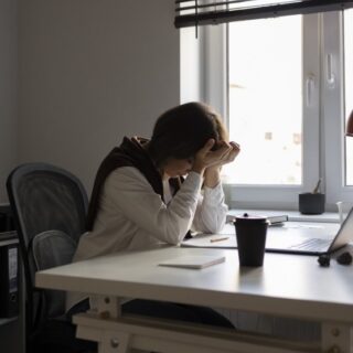 امرأة تجلس على كرسي ضمن المكتب وتضع يديها على رأسها وأمامها الحاسوب وأوراق على الطاولة