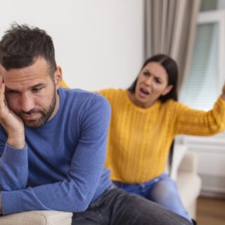 رجل يجلس على الأريكة مرتديا كنزة زرقاء اللون ويضع يده على خده وخلفه امرأة ترتدي كنزة صفراء اللون وتشير بيدها جانبا