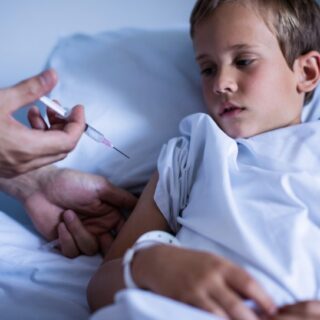 طفل ممدد على السرير في المستشفى وشخص يحقنه بإبره بكتفه