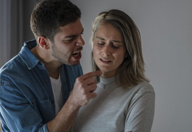 رجل يرتدي قميص أزرق ويصرخ بوجه امرأة مغمضة العينين ويرفع إصبعه بوجهها