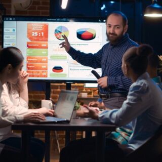 ثلاث نساء ورجل يجلسون على كراسي في المكتب ورجل يقف ويشير بيده لشاشة عليها بيانات