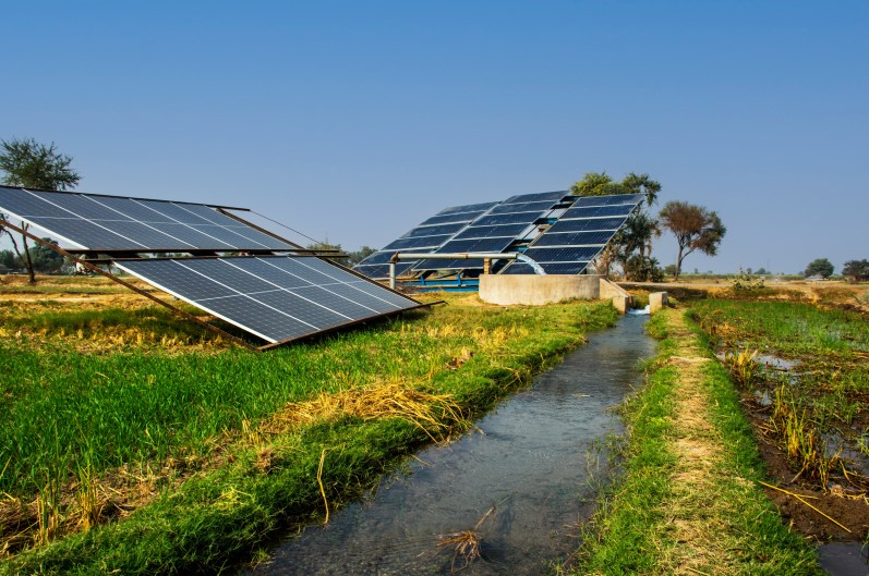 مجموعة من ألواح الطاقة الشمسية المتواجدة في حقل زراعي وبجانبها انبوب معدني تخرج منه المياه