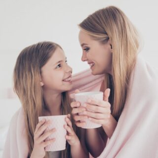 امرأة مبتسمة تمسك كوب بيدها وتنظر لفتاة تجلس بجانبها ويضعون غطاء على أكتافهم