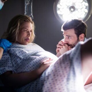 امرأة حامل في غرفة مظلمة ممسكة بيد زوجها ويدها الأخرى على بطنها وبجانبها ممرضة