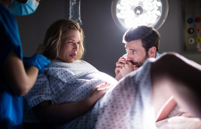 امرأة حامل في غرفة مظلمة ممسكة بيد زوجها ويدها الأخرى على بطنها وبجانبها ممرضة