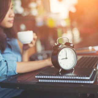 مراهقة تجلس على كرسي وتمسك كوب قهوة بيدها ويوجد بجانبها دفاتر ومنبه
