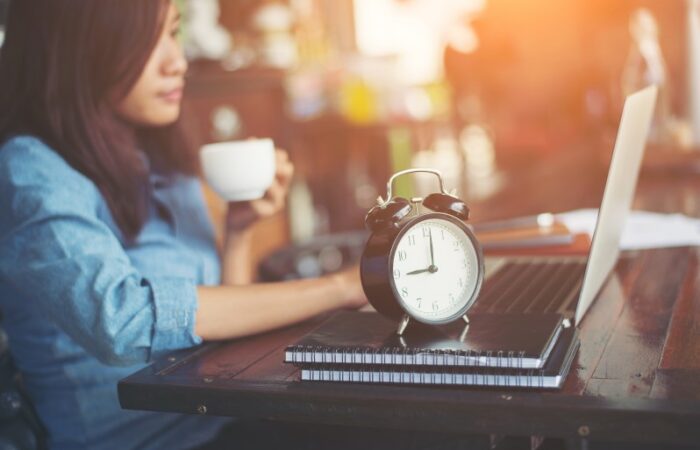 مراهقة تجلس على كرسي وتمسك كوب قهوة بيدها ويوجد بجانبها دفاتر ومنبه