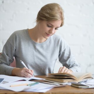 امرأة تمسك بقلم وتكتب على دفتر وتنظر لكتاب موجود أمامها على الطاولة