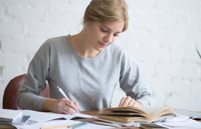 امرأة تمسك بقلم وتكتب على دفتر وتنظر لكتاب موجود أمامها على الطاولة