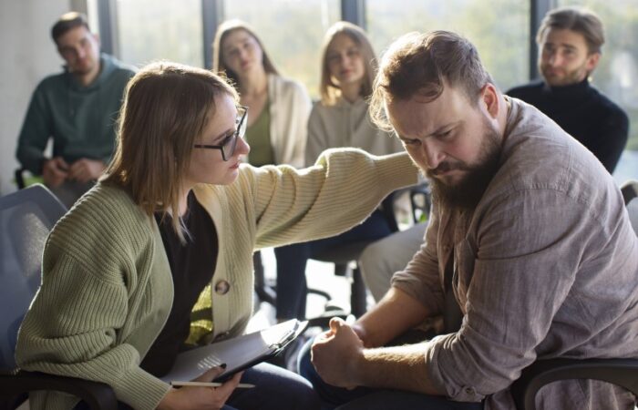 امرأة ترتدي نظارات وتمسك بقلم ودفتر وتضع يدها على كتف رجل ملتحي وحزين وخلفهم مجموعة من الأشخاص