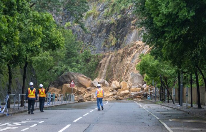 طريق جبلي مسدود بصخور كبيرة منهارة من جانب الطريق وثلاثة عمال طرق يقومون بمعاينة الموقع