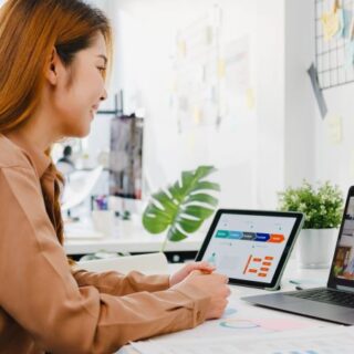 امرأة مبتسمة تنظر لشاشة اللابتوب وتتحدث مع أشخاص وبجانبها أيباد مرسوم على شاشته جدول بيانات