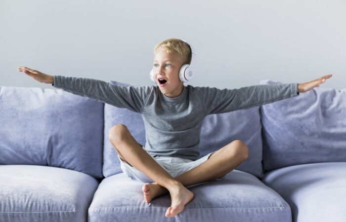 صبي يجلس على الأريكة ويضع سماعات بيضاء على أذنيه ويفتح ذراعيه جانبا